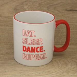 May include: White ceramic mug with a red rim and handle. The mug has red text that reads "Eat. Sleep. Dance. Repeat."
