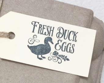 May include: A set of four brown paper gift tags with a black silhouette of a duck and the text "Fresh Duck & Eggs". The tags are tied with twine and are sitting on top of a brown cardboard egg carton.
