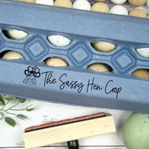 Puede incluir: Una huevera azul llena de huevos moteados, con el texto "The Sassy Hen Coop" impreso en el lateral. Tambi&eacute;n hay un sello de madera y huevos decorativos, creando una est&eacute;tica r&uacute;stica y fresca de granja.