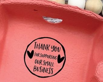 Op de afbeelding: Een roze kartonnen eierdoos met zes blauwe eieren erin. De doos heeft een zwart-witte sticker met de tekst "Bedankt voor het steunen van ons kleine bedrijf" met twee harten.