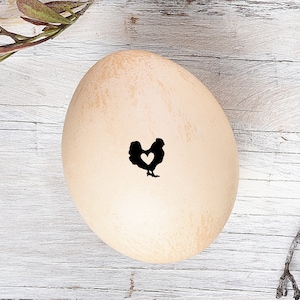 May include: Close-up view of a light beige speckled egg with a small black silhouette of a chicken imprinted on its surface. The chicken design incorporates a small heart shape within its body. The egg rests on a white wooden surface, with a branch featuring green leaves subtly visible in the upper left corner. The overall image suggests a rustic, farmhouse aesthetic, possibly related to Easter or spring-themed decorations.