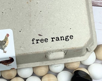 May include: A brown cardboard egg carton with a white sticker that says "free range" and a picture of a chicken. A wooden stamp with the text "free range" is on a white surface with wooden beads.
