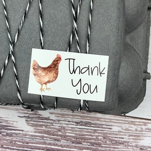 May include: A gray egg carton tied with black and white twine. A small white sticker with a brown chicken illustration and the text 