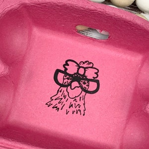May include: Pink six-egg carton with a black graphic of a chicken wearing glasses.  The carton is made of cardboard and contains six white eggs.  The chicken illustration is simple and features a bow on its head. The carton is shown against a white wooden background with a beaded garland.