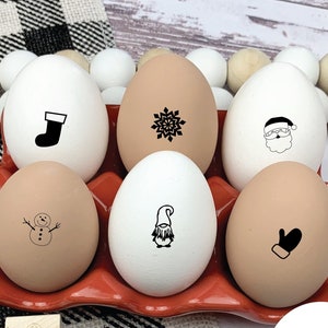 May include: Six white eggs with black Christmas designs in an egg carton. The designs include a stocking, snowflake, Santa Claus, snowman, gnome, and mitten. There are also six wooden stamps with the same designs. The stamps are arranged on a black and white checkered surface. A white circle with the text 