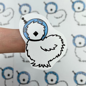 May include: Nine white chick stickers with blue astronaut helmets. The stickers are on a white sheet of paper with a pink border. The stickers are arranged in three rows of three.