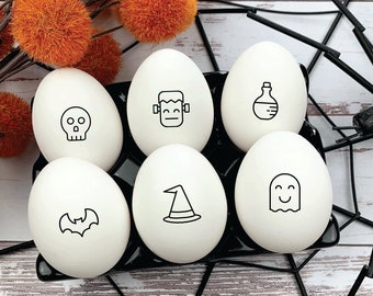 May include: Six white eggs with black Halloween-themed illustrations. The eggs are arranged in a black egg carton. The illustrations include a skull, a bat, a witch's hat, a ghost, a potion bottle, and a monster.