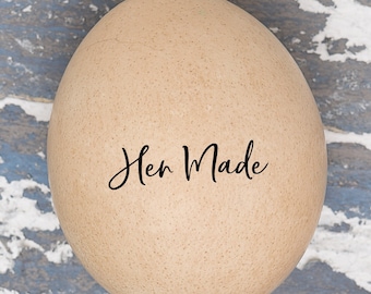 May include: A brown egg with the text "Hen Made" written on it. The egg is sitting on a blue and white wooden surface.