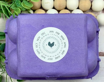 May include: A purple egg carton with a white sticker featuring a calendar with a chicken silhouette. The calendar shows the months of the year and the days of the week. The sticker has a wreath design around the calendar.