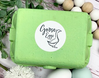 May include: A green cardboard egg carton with a white sticker that says "Goose Eggs" and a black and white illustration of a goose. There are three white eggs and one light blue egg on a white wooden surface.