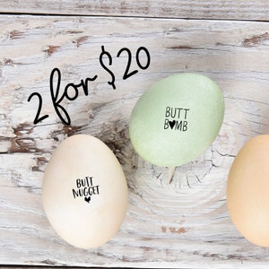 May include: Two eggs, one green and one beige, on a white wooden surface. The green egg has the text 