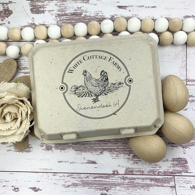 May include: A beige egg carton with the text "White Cottage Farms" and a hen illustration. Three wooden eggs are next to the carton. Decorative wooden beads and a burlap flower are in the background.