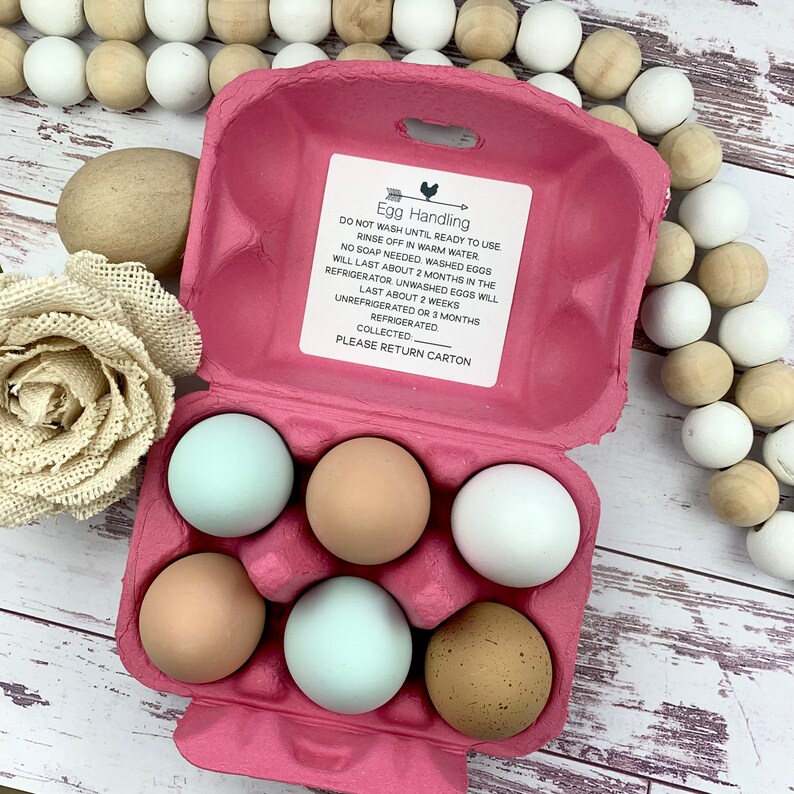 Puede incluir: Un cart&oacute;n de huevos de cart&oacute;n rosa con seis huevos marrones, blancos y azules en su interior. El cart&oacute;n tiene una etiqueta blanca con el texto "Egg Handling" e instrucciones para manipular y almacenar los huevos.