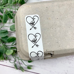 May include: Three white stickers with black duck and arrow designs on a gray egg carton. Each sticker has a duck inside a heart with an arrow through it.