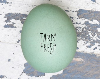 May include: A single, light green egg with the words "FARM FRESH" written on it. The egg is sitting on a white and blue painted wooden surface.
