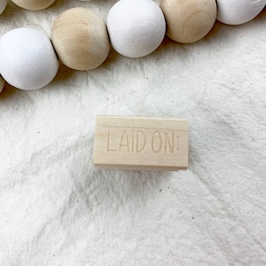 May include: A wooden block stamp with the text "LAID ON:" engraved on the surface. The stamp is sitting on a white fabric background.