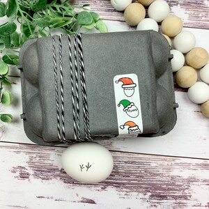 May include: A gray egg carton with a white and black twine tied around it. A sticker with three cartoon eggs wearing different hats is on the side of the carton. A white egg with a drawing of a chicken foot on it is in front of the carton.