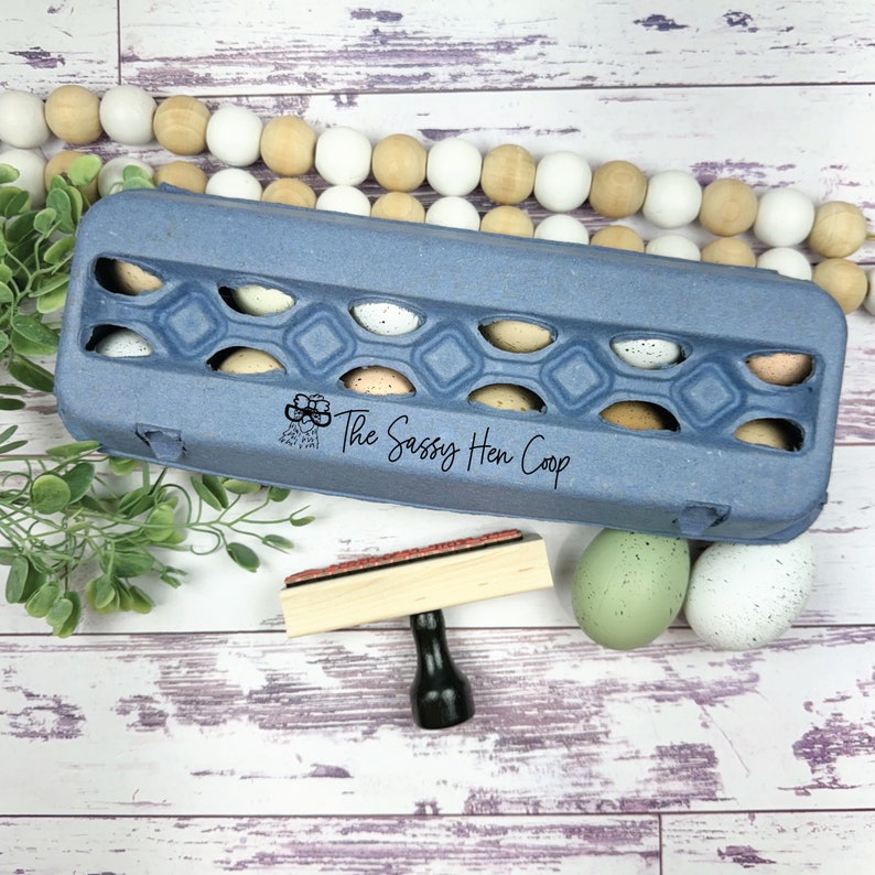Puede incluir: Una huevera azul llena de huevos moteados, con el texto "The Sassy Hen Coop" impreso en el lateral. Tambi&eacute;n hay un sello de madera y huevos decorativos, creando una est&eacute;tica r&uacute;stica y fresca de granja.