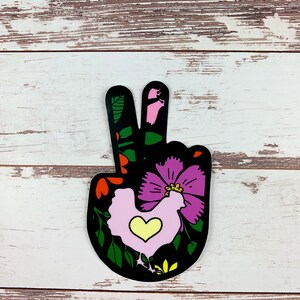 May include: A black peace sign sticker with a pink and purple flower and a pink chicken with a yellow heart in the centre. The sticker has a green, black, and red floral design.