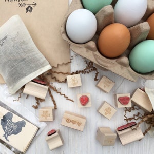 Puede incluir: Un conjunto de sellos de madera con varios dise&ntilde;os, incluyendo corazones, signos de paz, pollos y huevos. Los sellos est&aacute;n dispuestos sobre una superficie blanca con virutas de papel marr&oacute;n y una caja de cart&oacute;n para huevos con varios huevos.