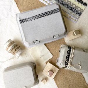 Puede incluir: Vista a&eacute;rea de cajas de huevos grises adornadas con cinta adhesiva y lazos con dise&ntilde;os en blanco y negro. Suministros de artesan&iacute;a: sello de madera, peque&ntilde;a bolsa de tela y tiras de papel decorativas. Tema hecho a mano.