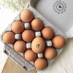 Puede incluir: Una docena de huevos marrones en una caja de cart&oacute;n gris. Un huevo est&aacute; estampado con la palabra "LOVE" y un peque&ntilde;o dibujo de un pollo. La caja tiene un sello circular con meses y fechas.
