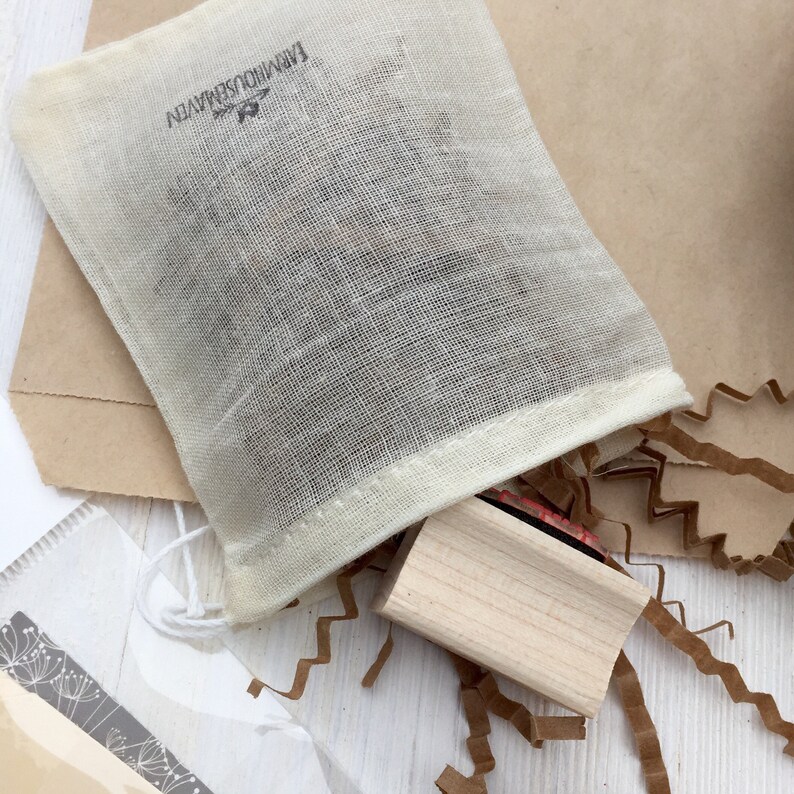 K&ouml;nnte beinhalten: Ein wei&szlig;er Musselinbeutel mit einem schwarzen Stempel, auf dem "Handmade with Love" steht, und einem Holzstempel mit einem roten Stempelkissen. Der Beutel liegt auf einer wei&szlig;en Oberfl&auml;che mit braunen Papierfetzen.
