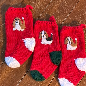 Puede incluir: Tres calcetines de Navidad de punto rojo con puños blancos y talones verdes. Cada calcetín tiene una aplicación de fieltro de un perro beagle marrón y blanco.
