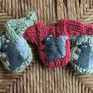 May include: Three knitted ornaments shaped like sweaters with a gray felt squirrel appliqué on each. The sweaters are green, red, and green.