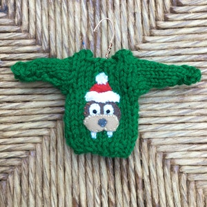 May include: A green knitted sweater ornament with a brown felt animal wearing a red Santa hat. The ornament is hanging from a gold wire hanger.