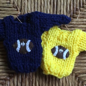 Op de afbeelding: Twee gebreide truien met voetbalmotieven. De ene trui is donkerblauw en de andere is geel.