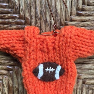 Op de afbeelding: Oranje gebreide trui met een bruine vilt voetbal applicatie. De trui hangt aan een draadhangertje.
