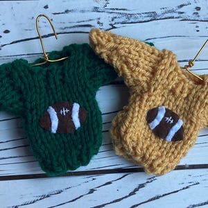 Op de afbeelding: Twee miniatuur gebreide truien met een voetbalontwerp. De ene trui is groen en de andere is geel.