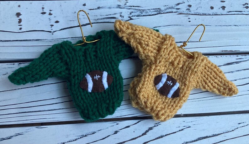 Op de afbeelding: Twee miniatuur gebreide truien met een voetbalontwerp. De ene trui is groen en de andere is geel.