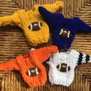 Op de afbeelding: Vier miniatuur gebreide truien met voetbalmotieven. De truien zijn geel, blauw, oranje en wit met zwarte strepen. Elke trui heeft een bruine viltvoetbal met witte stiksels.