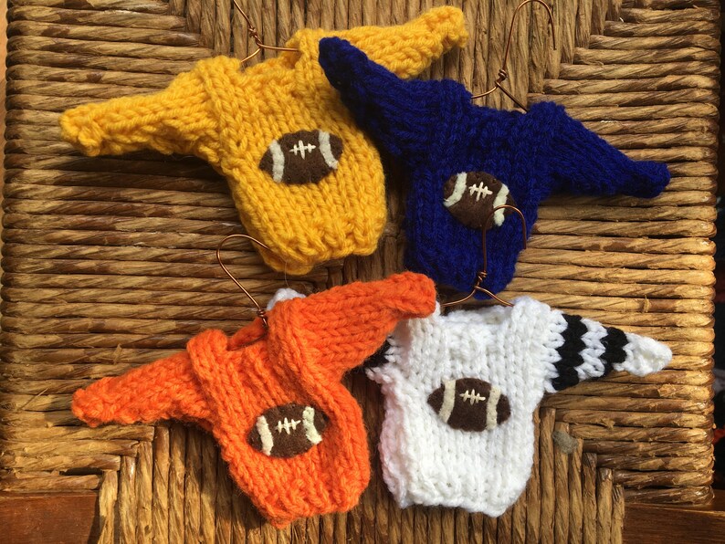 Op de afbeelding: Vier miniatuur gebreide truien met voetbalmotieven. De truien zijn geel, blauw, oranje en wit met zwarte strepen. Elke trui heeft een bruine viltvoetbal met witte stiksels.