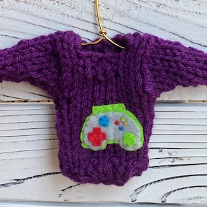 以下が含まれることがあります： 緑、グレー、赤のフェルトでできたビデオゲームコントローラーのアップリケが付いた紫色のニットセーター