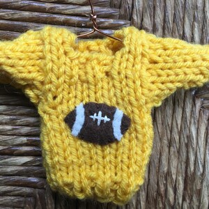 Op de afbeelding: Een gele gebreide trui met een bruine vilt voetbal applicatie. De trui hangt aan een hanger.