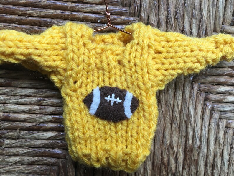 Op de afbeelding: Een gele gebreide trui met een bruine vilt voetbal applicatie. De trui hangt aan een hanger.