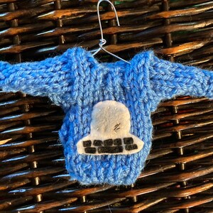 Pode incluir: Um enfeite de suéter azul de tricô com uma aplicação de nave espacial de feltro branco. O enfeite está pendurado em um cabide de arame.