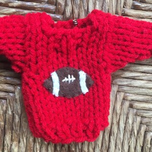 Op de afbeelding: Een rode gebreide trui met een bruin en wit voetbalontwerp op de voorkant.