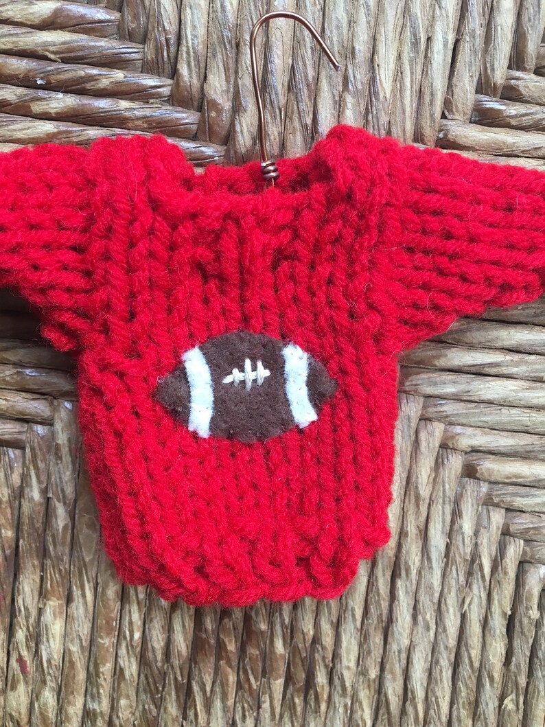 Op de afbeelding: Een rode gebreide trui met een bruin en wit voetbalontwerp op de voorkant.