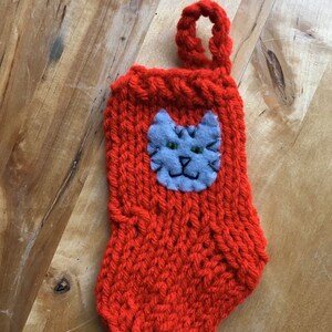 May include: A red knitted stocking with a gray felt cat face appliqué. The cat has green eyes and a stitched smile.