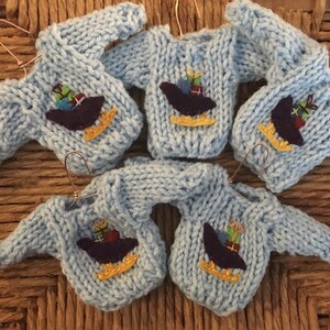 May include: Five miniature blue knitted sweaters with a festive design of a sled carrying presents. Each sweater has a gold wire hanger.