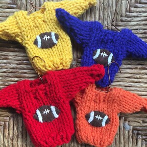 Op de afbeelding: Vier miniatuur gebreide truien in geel, blauw, rood en oranje, elk met een bruine vilt voetbal applicatie.
