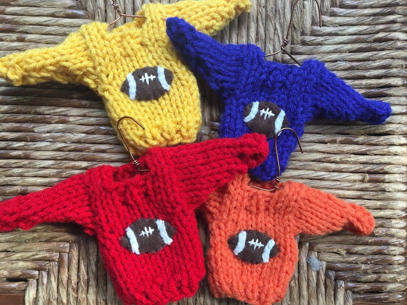 Op de afbeelding: Vier miniatuur gebreide truien in geel, blauw, rood en oranje, elk met een bruine vilt voetbal applicatie.