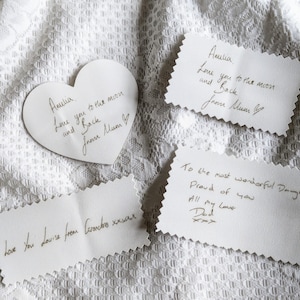 May include: Close-up of white paper notes with handwritten messages, including a heart-shaped note. The messages say "Love you to the moon and back" and "To the most wonderful Daughter". The notes are on a white lace surface.