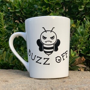 May include: White ceramic mug with a black and white illustration of a bee with an angry face. The text "Buzz Off" is printed below the bee.