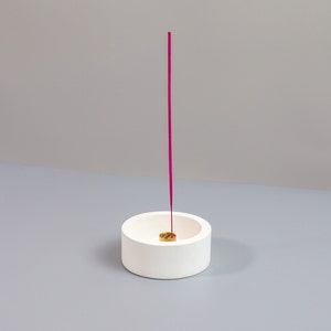 May include: A white, cylindrical incense holder with a small, round, gold-colored insert. A tall, thin, magenta incense stick is inserted into the holder, standing upright. The background is a solid, light gray.