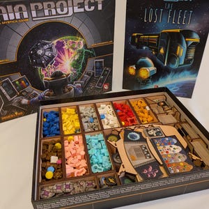 May include: A Gaia Project board game box with a wooden organizer insert containing various colored plastic game pieces, including blue, yellow, gray, white, red, orange, brown, pink, and light blue tokens, alongside game boards and cards.